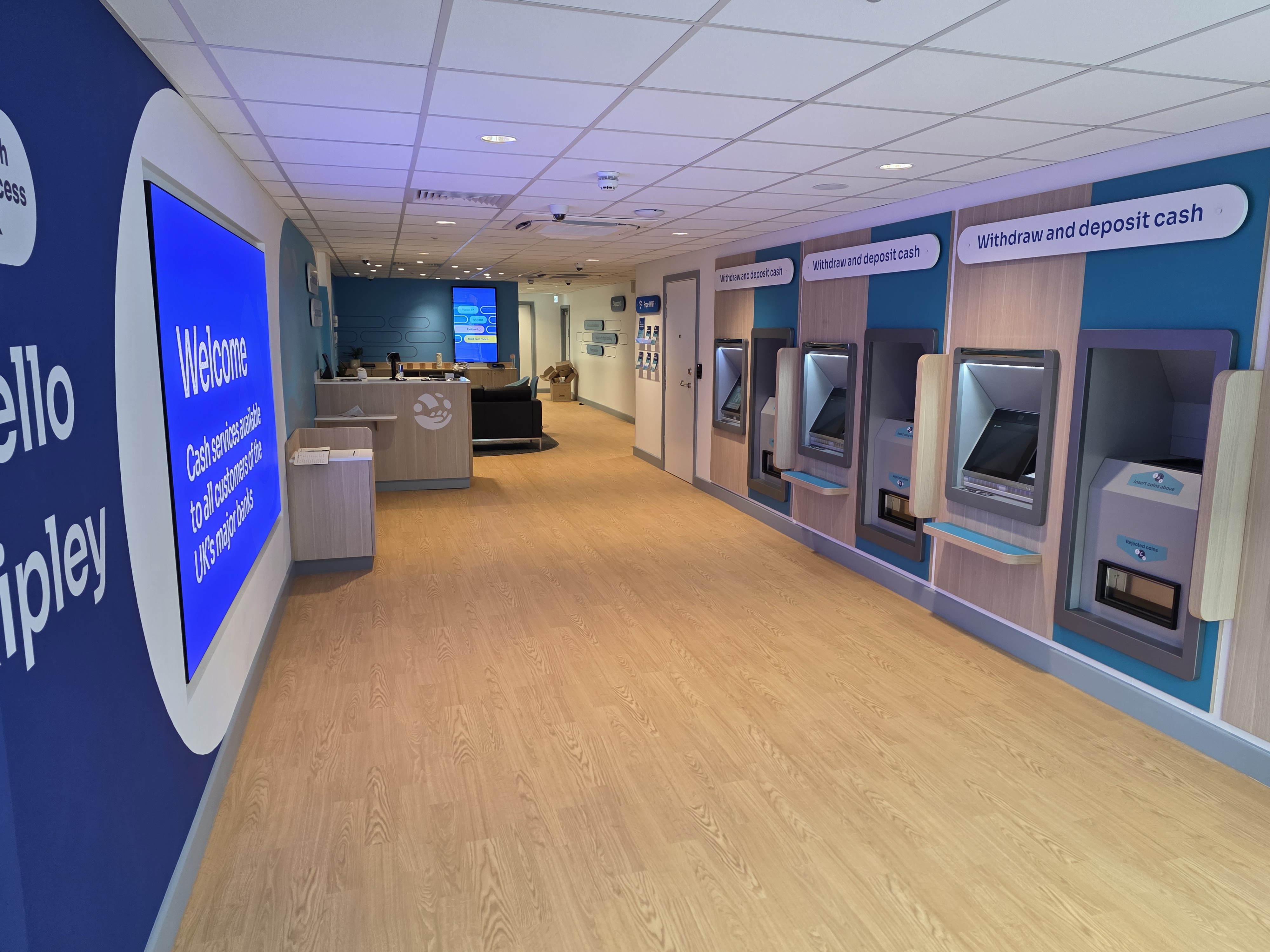 Accessible banking hub interior with a row of self-service cash machines on the right, a welcome sign on the left wall, and a staffed counter and seating area toward the back in a clean, modern space.