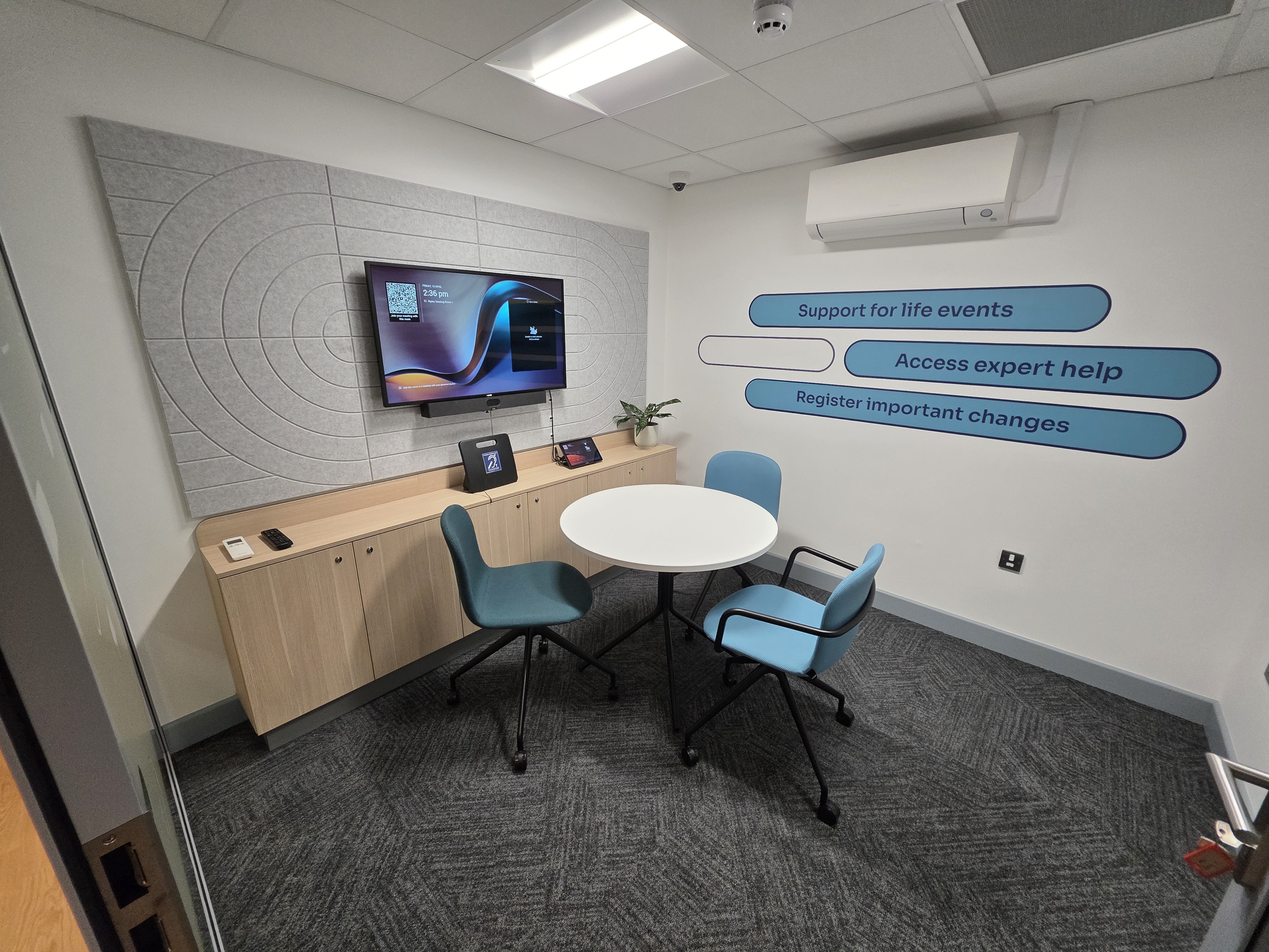 Private consultation room, featuring a round table with three chairs, a wall-mounted screen above a wooden cabinet, and signage highlighting support for life events, expert help, and registering important changes.